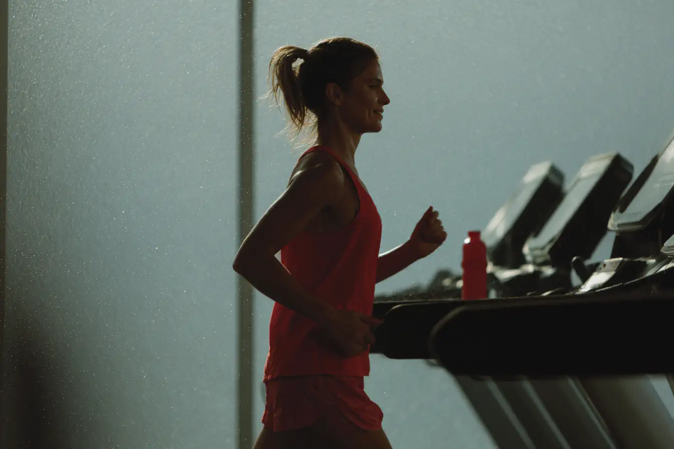 Casasports Eine Frau in Sportkleidung joggt auf einem Laufband in einer Halle. Sie ist im Profil zu sehen, beleuchtet von sanftem Licht, und hat eine Wasserflasche vor sich auf dem Laufband stehen. Fitnessstudio Oer-Erckenschwick