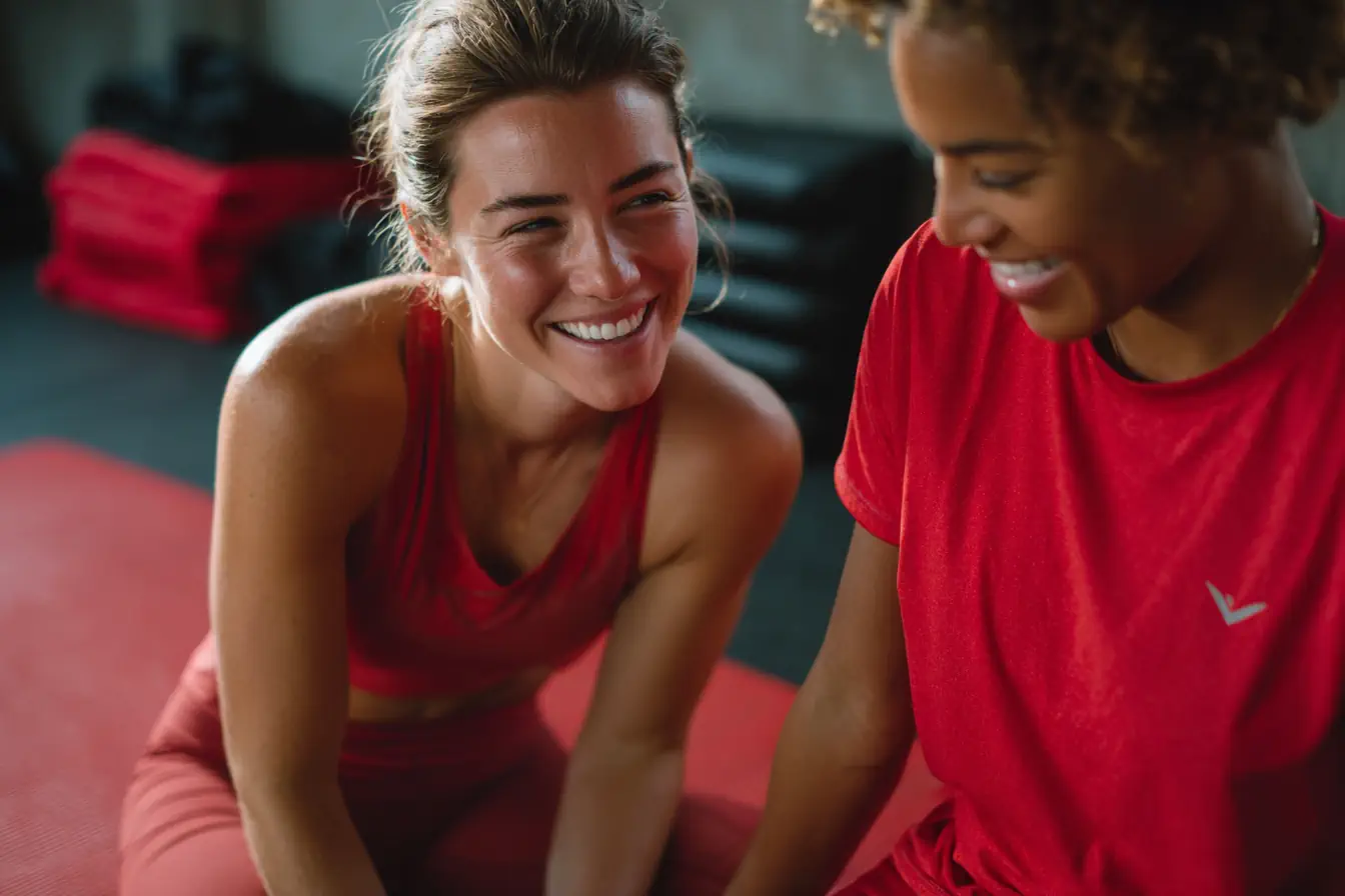 Casasports Zwei Frauen in roter Sportkleidung sitzen auf einer Matte in einem Fitnessstudio, lächeln und führen ein freundliches Gespräch nach einem Workout. Der Hintergrund ist leicht unscharf, die Fitnessgeräte sind sichtbar. Fitnessstudio Oer-Erckenschwick
